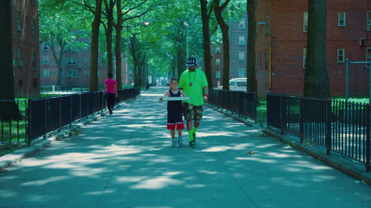 Red Hook Summer (2012) movie still: fullbody — A paved path between two red brick apartment complexes is shaded by large trees. An adult man and a…