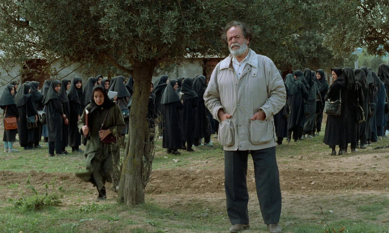 Through The Olive Trees (1994) movie still: fullbody — A rural outdoor space with a line of women in black clothing in background. An older man…