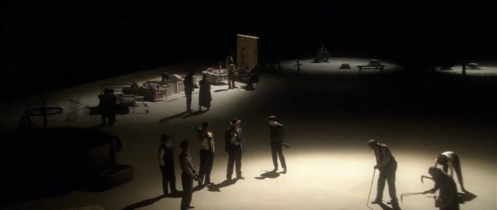 Manderlay (2005) movie still: wide — A dark, empty soundstage contains disconnected islands of furniture and props under pools of light. Several men…