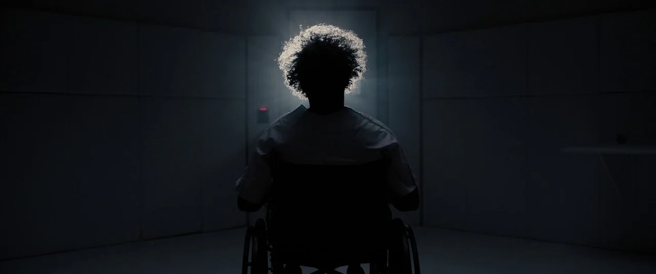 Glass (2019) movie still: medium — A dark, clinical room features plain white wall panels and a faint red indicator light. A person with curly hair…