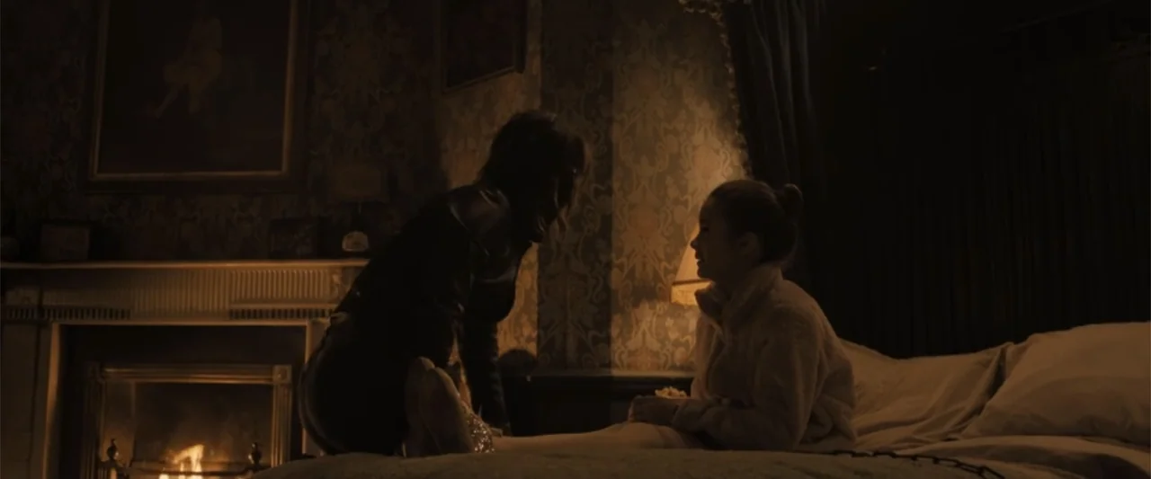 Abigail (2024) movie still: medium — A dimly lit bedroom features patterned wallpaper and a fireplace glowing in the background. A woman leans…