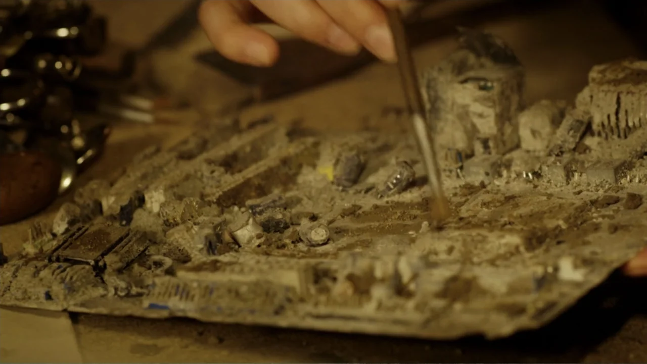 Mortal Engines (2018) movie still: closeup — A dusty, damaged circuit board sits on a workbench in a cluttered interior space. A hand uses a…