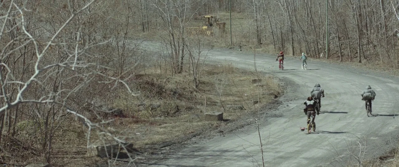 Turbo Kid (2015) movie still: wide — A rural dirt path winds through a sparse landscape of bare winter trees. Four individuals ride bicycles away…