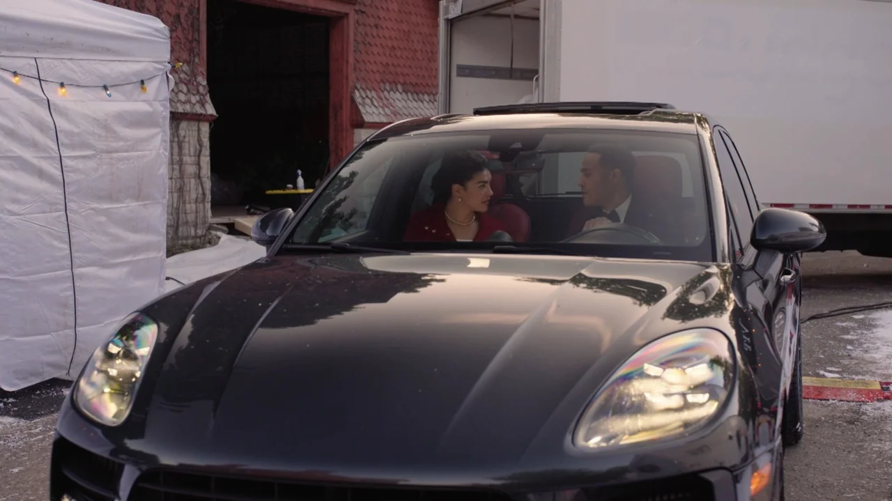 Designing Christmas (2022) movie still: medium — A dark luxury car sits in a parking lot next to a white tent. A man and woman sit inside the…