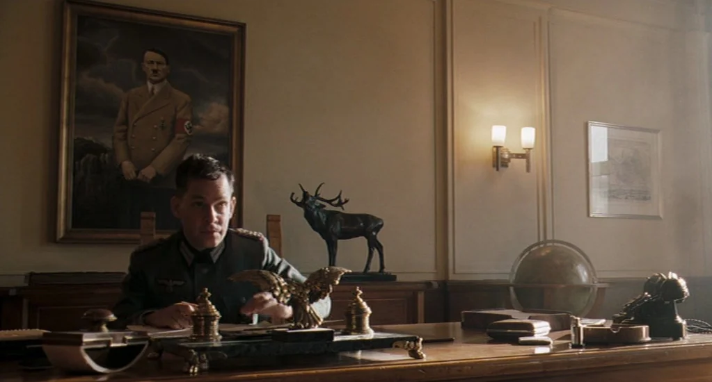 Valkyrie (2008) movie still: medium — A German military officer sits at a desk in a wood-paneled room. Behind him hangs a portrait of Adolf Hitler.