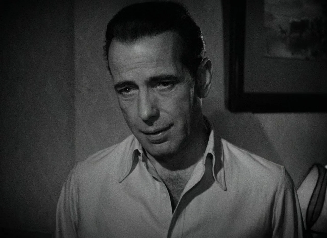 Key Largo (1948) movie still: closeup — A dimly lit indoor space with a picture frame visible on the wall. A man in a light-colored shirt gazes…