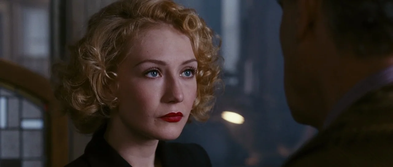 Black Book (2006) movie still: closeup — A dimly lit room with a window visible in the background shadows. A woman with curled blonde hair looks…