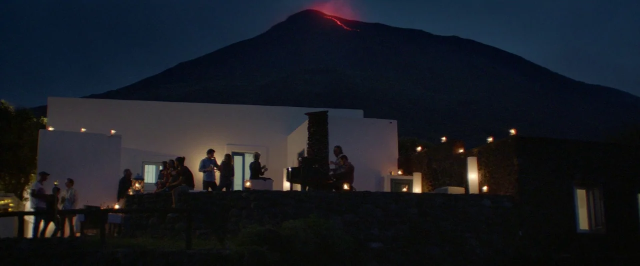 Sibyl (2019) movie still: wide — A white house sits on a stone terrace at the base of a dark volcano. A group of people gathers on the patio while…