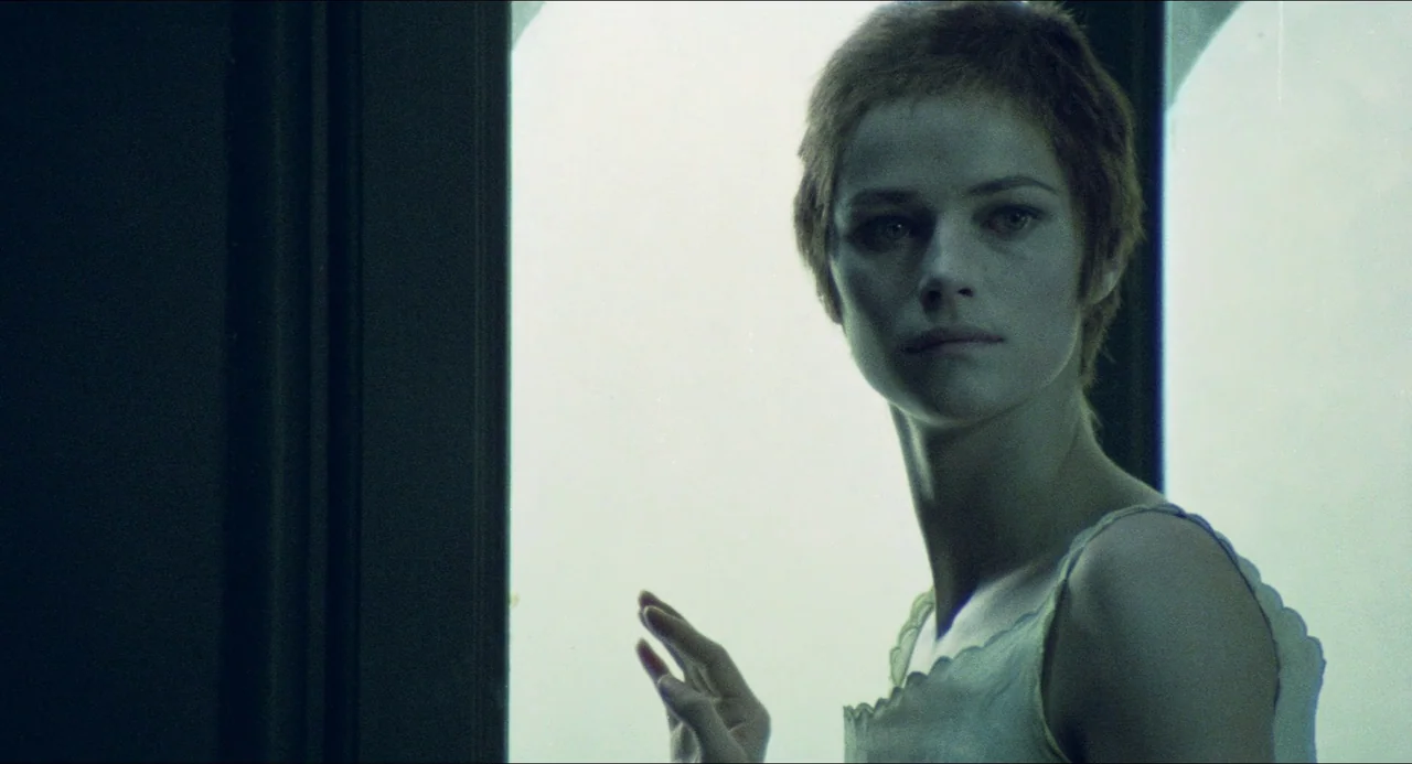 The Night Porter (1974) movie still: closeup — A woman stands indoors near a bright window with a clear pane of glass. She turns her head toward the…