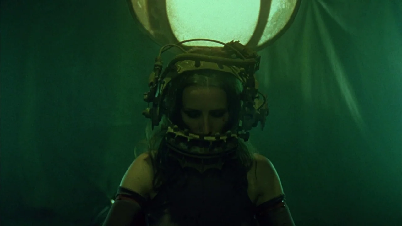 Saw movie still: medium — A dimly lit enclosed space with a dark backdrop and green ambient light. A woman is positioned frontally while wearing a…