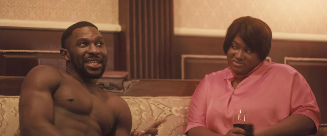 Casa De Novia (2024) movie still: medium — A refined interior setting features beige patterned walls and an ornate brown sofa. A muscular shirtless…