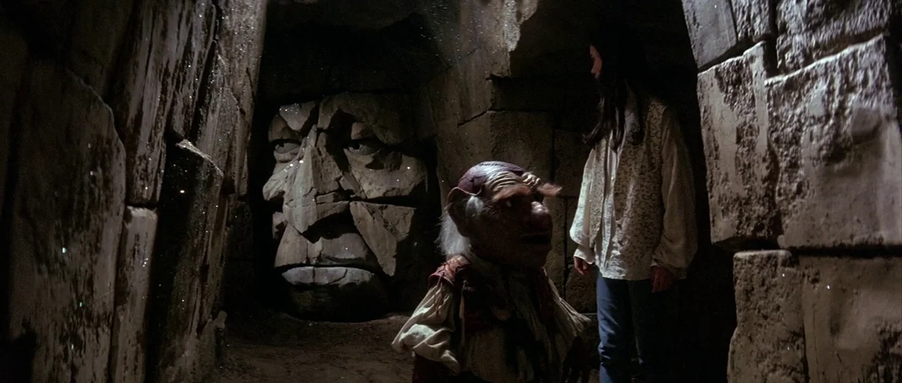 Labyrinth (1986) movie still: medium — A dark, narrow passageway made of rough-hewn stone blocks under low light conditions. A small goblin-like…