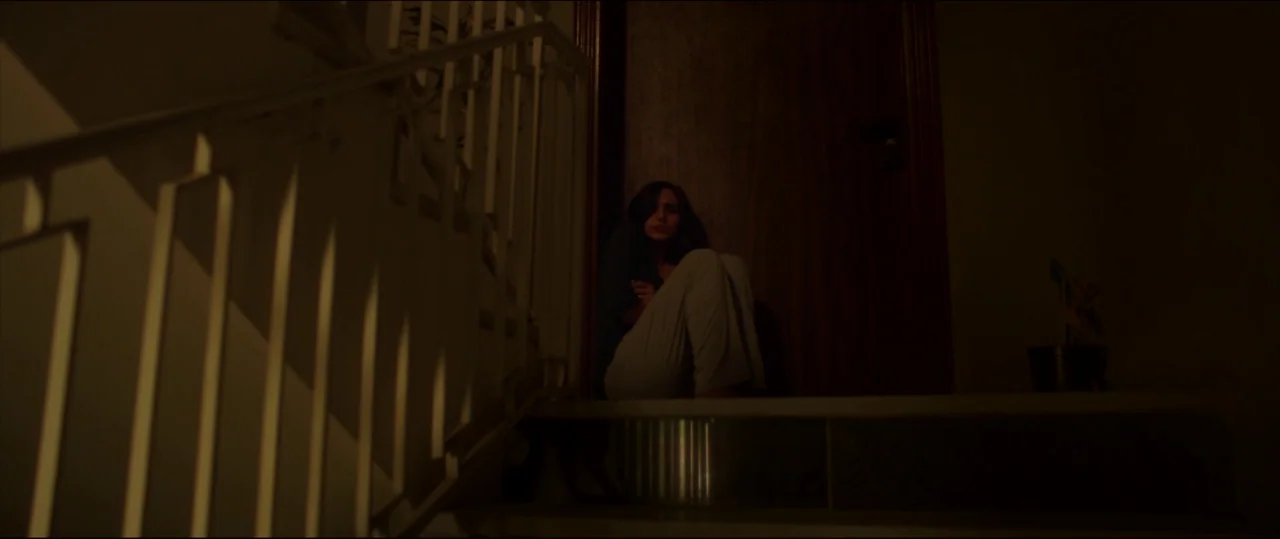 Under The Shadow (2016) movie still: wide — A dimly lit indoor staircase features a white metal railing and dark wooden door. A woman sits on the…