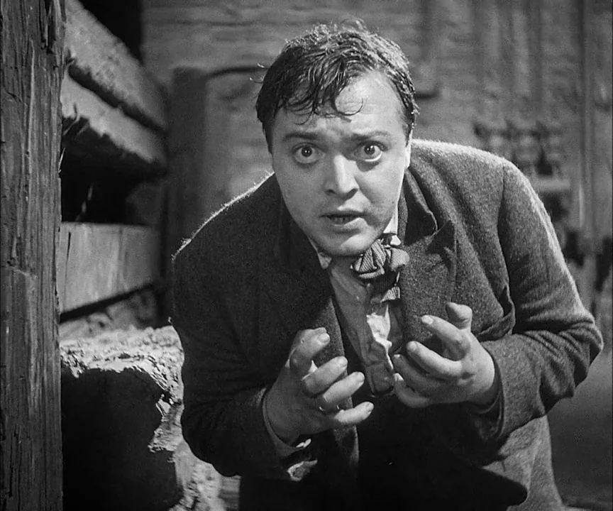 M (1931) movie still: closeup — A man stands in front of a dark, textured brick wall at night. He is reacting to something unseen while holding his…