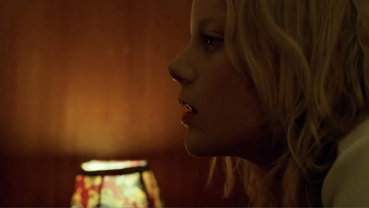 Emily (1976) movie still: closeup — A dimly lit room features a patterned lampshade placed against dark brown wooden paneling. A young woman with…