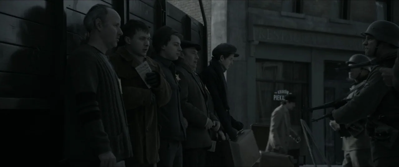 Bau, Artist at War (2025) movie still: medium — A mid-1940s city street with brick architecture and storefronts in the background. Several civilians…