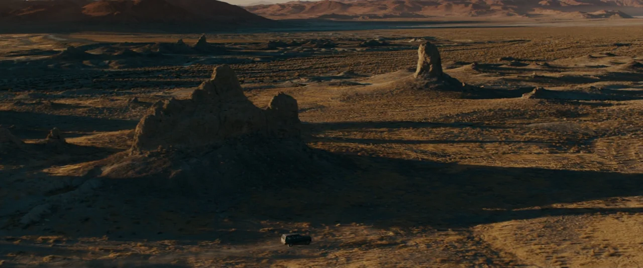Encounter (2018) movie still: establishing — An arid, vast desert landscape features jagged rock formations under a clear sky. A lone dark vehicle…