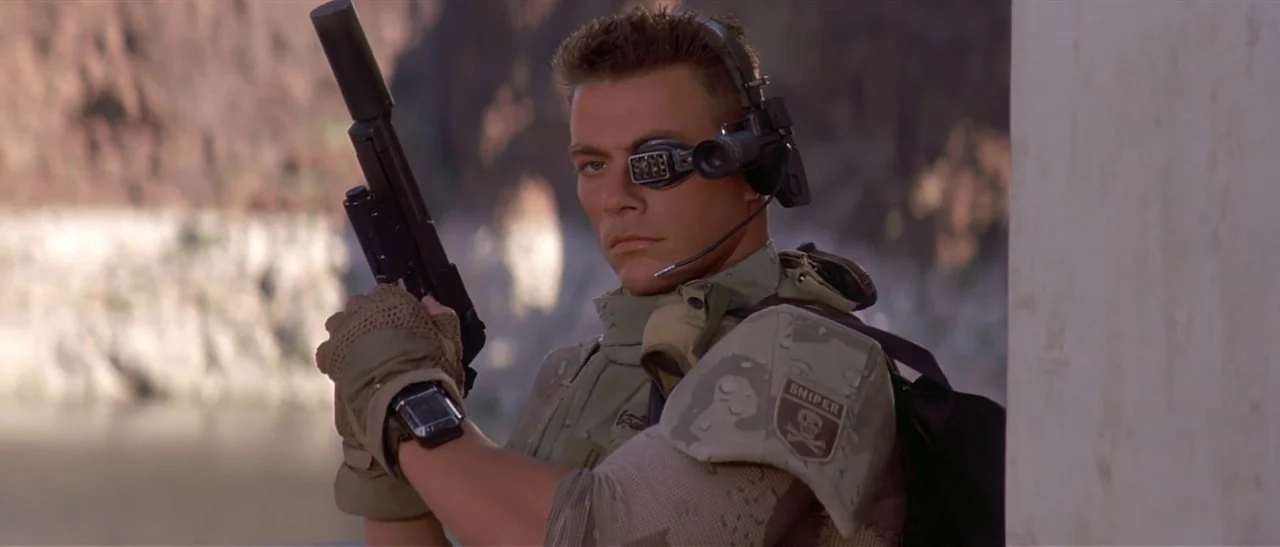 Universal Soldier (1992) movie still: medium — A soldier stands outdoors in a rocky environment wearing desert tactical gear and equipment. He holds…