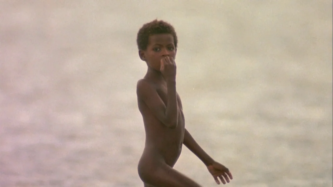 Powaqqatsi (1988) movie still: fullbody — A young boy stands in shallow water against a bright, hazy, light-colored background. The child is…