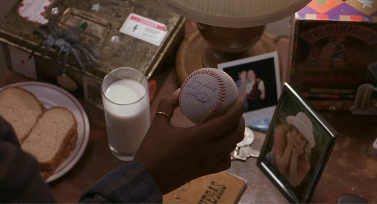 Bogus (1996) movie still: closeup — A cluttered wooden tabletop contains a glass of milk, a baseball, and various photographs. A hand enters the…
