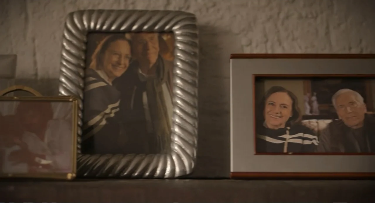 La posada (2023) movie still: closeup — A wooden shelf holds several framed photographs featuring the same couple in each picture. The silver and…