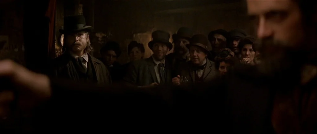 The Prestige (2006) movie still: medium — A dimly lit 19th-century pub interior filled with a crowd of men in hats. The men watch a central figure…