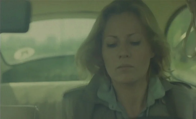 No End (1985) movie still: closeup — The interior of a 1970s car cabin shows a view through the windshield. A woman sits in the driver's seat and…