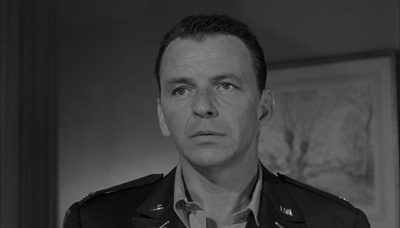 The Manchurian Candidate (1962) movie still: closeup — An indoor office setting with a framed picture visible on the wall behind. A man in a…