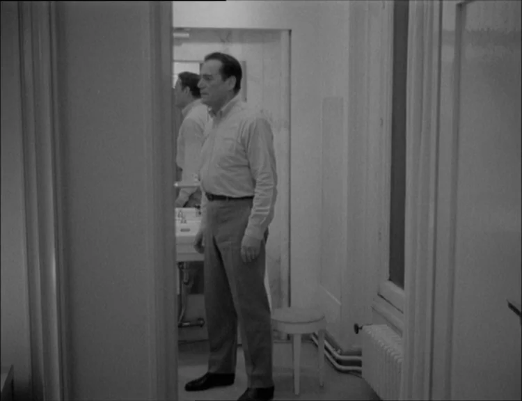 Alphaville (1965) movie still: fullbody — A sterile bathroom interior features a vanity mirror and a small white stool. A middle-aged man stands in…