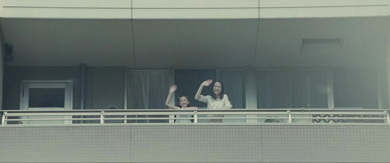 The World of Kanako (2014) movie still: wide — A modern concrete apartment building features a wide balcony with horizontal metal railings. A woman…