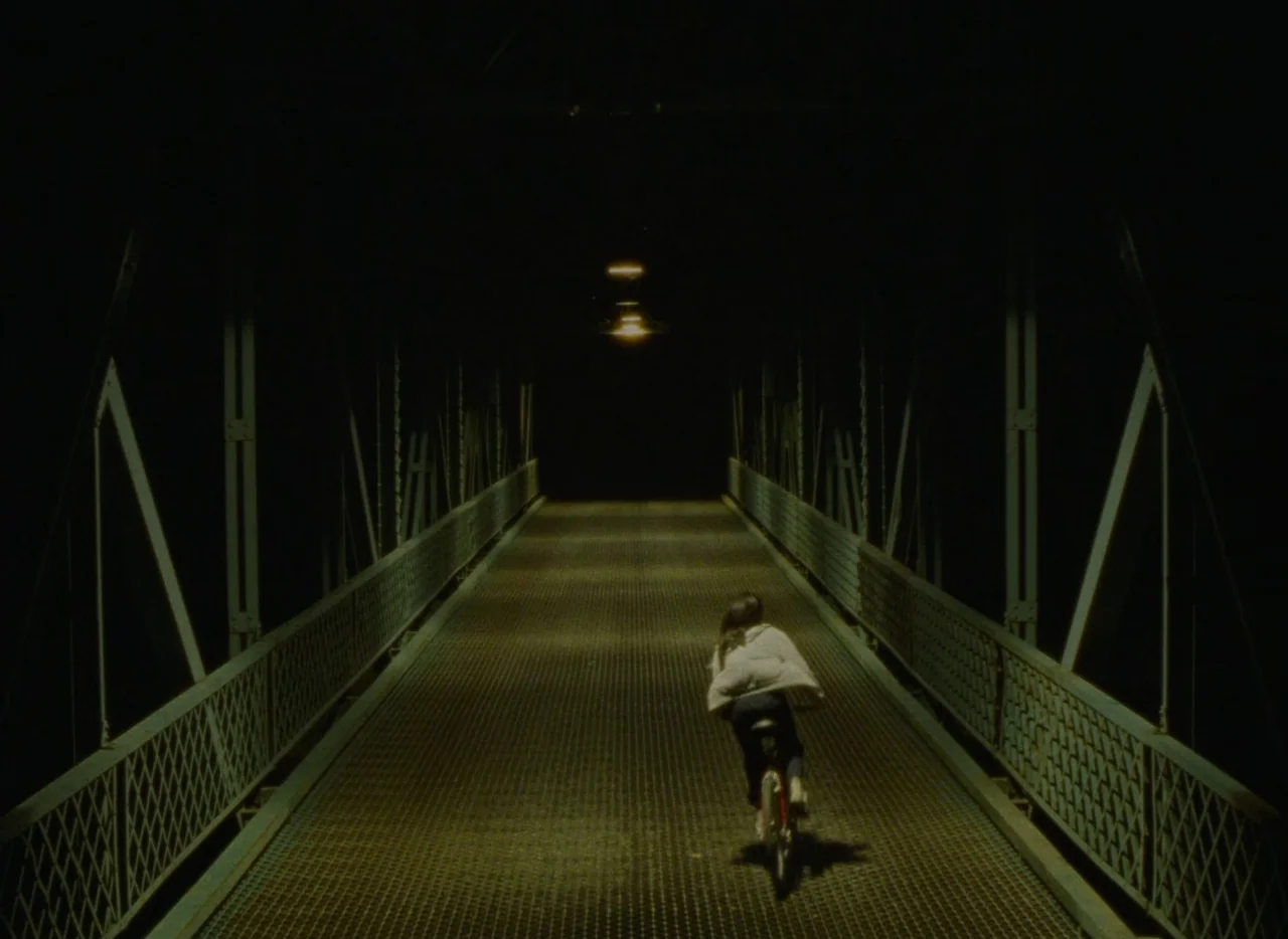 Falcon Lake (2022) movie still: wide — A dark pedestrian bridge constructed of metal trusses spans a black void. A solitary child on a bicycle rides…