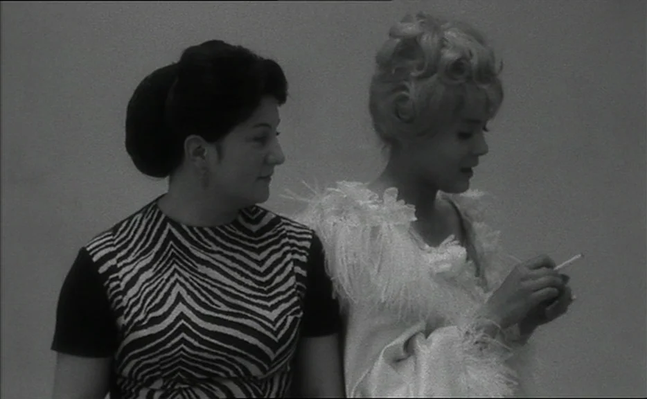 Cléo from 5 to 7 movie still: medium — Two women stand against a plain, light-colored background. The woman on the right holds a lit cigarette while…