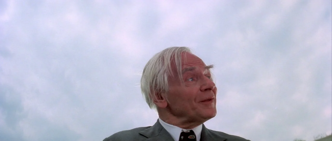 Suspiria (1977) movie still: closeup — An elderly man in a suit stands outdoors against a bright, overcast sky. The subject gazes upward toward an…