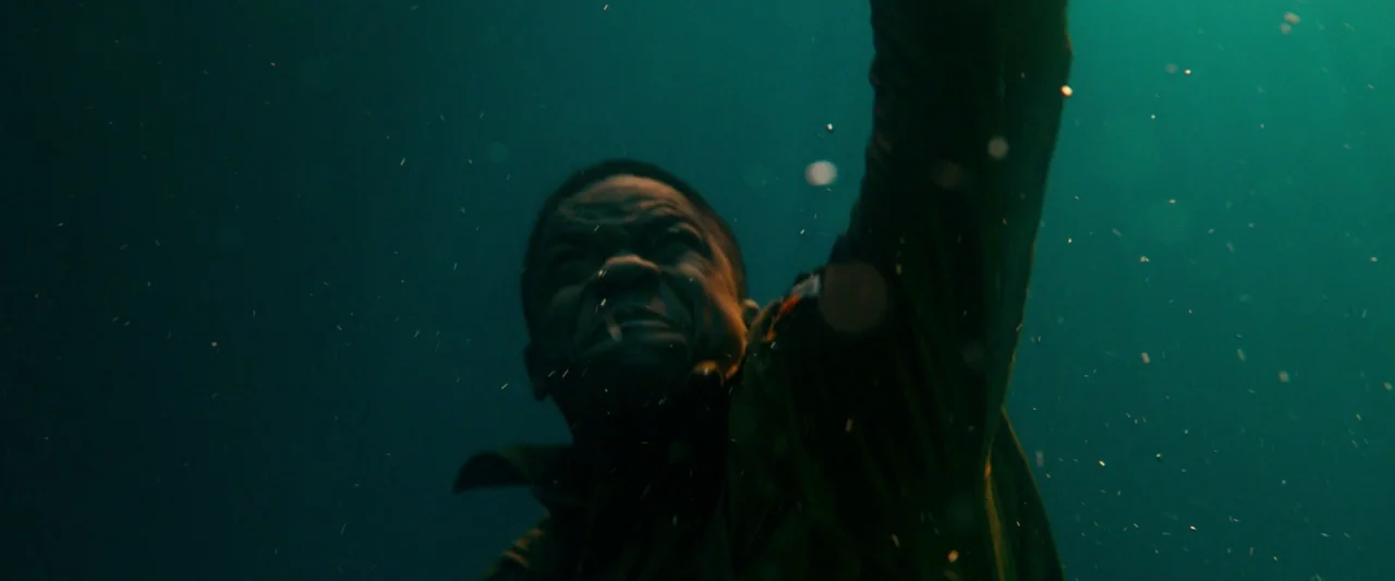 Overlord (2018) movie still: closeup — A dark, deep underwater space filled with floating particulates and filtered green light. A man strains as he…