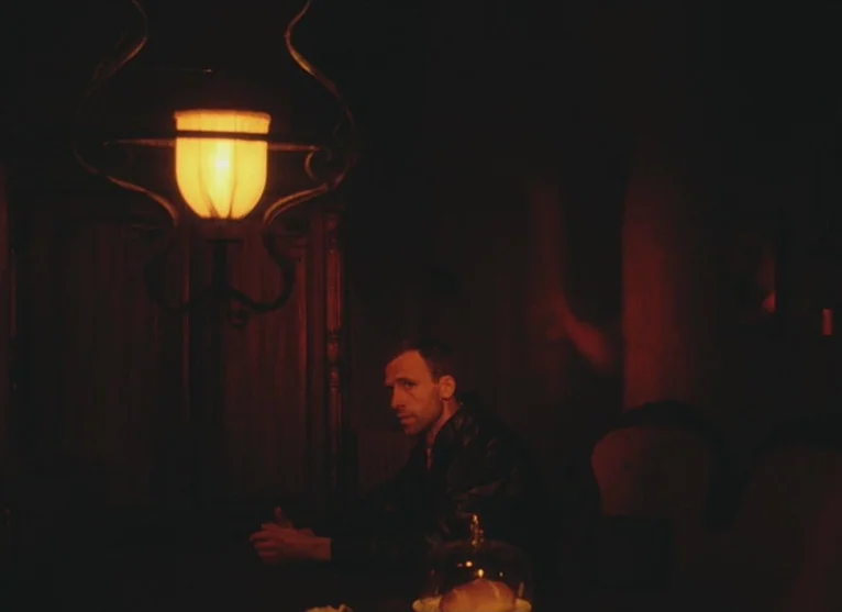 A Visitor to a Museum (1989) movie still: medium — A dimly lit room with dark wooden paneling and minimal visible furniture. A man sits alone at a…