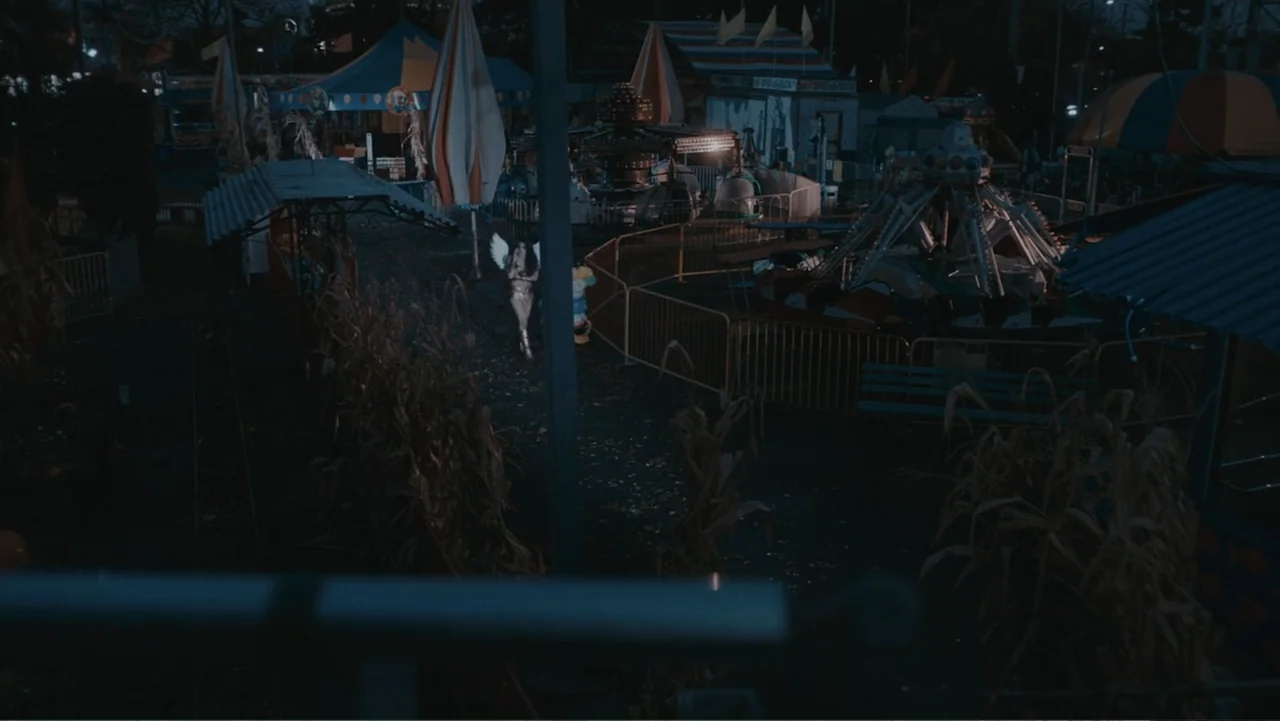 Terrifier 2 (2022) movie still: wide — A nighttime fairground with various rides and dry corn stalks lining the walkways. A person with white wings…