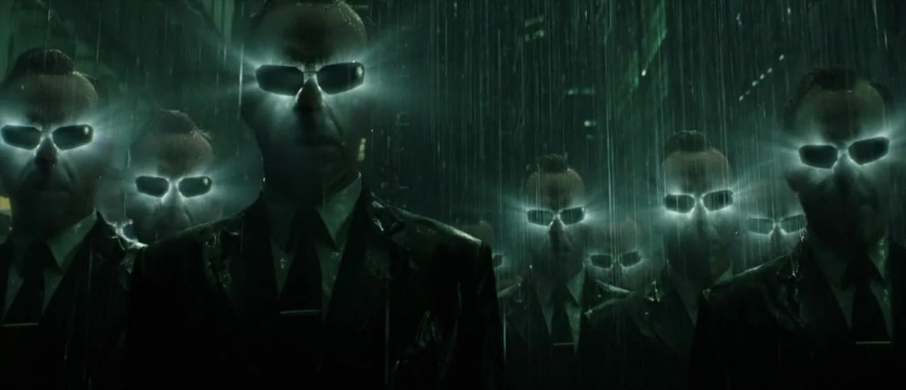 The Matrix Revolutions (2003) movie still: medium — A group of identical men in black suits stands in a rain-slicked city street. Each man wears…