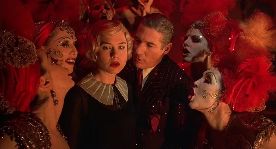 Chicago (2002) movie still: medium — A crowded cabaret setting features performers with elaborate feathered headpieces and dramatic stage makeup. A…