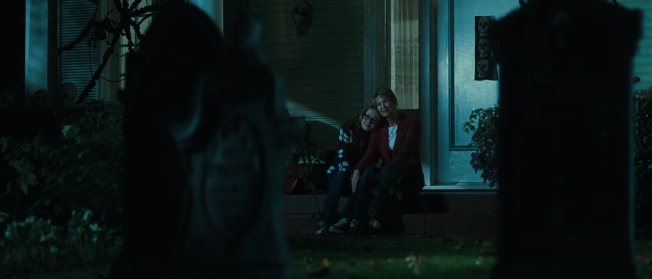 Halloween (2007) movie still: wide — A dark, blue-toned front porch decorated with Halloween skulls on the lawn. Two women sit on the concrete porch…