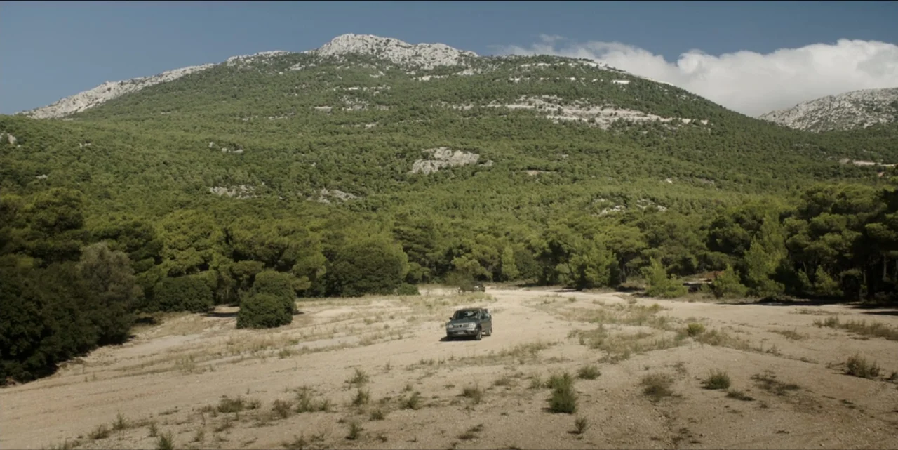 The Hunted (2024) movie still: establishing — A wide valley sits between a large, tree-covered mountain and sparse dry ground. A small SUV is parked…