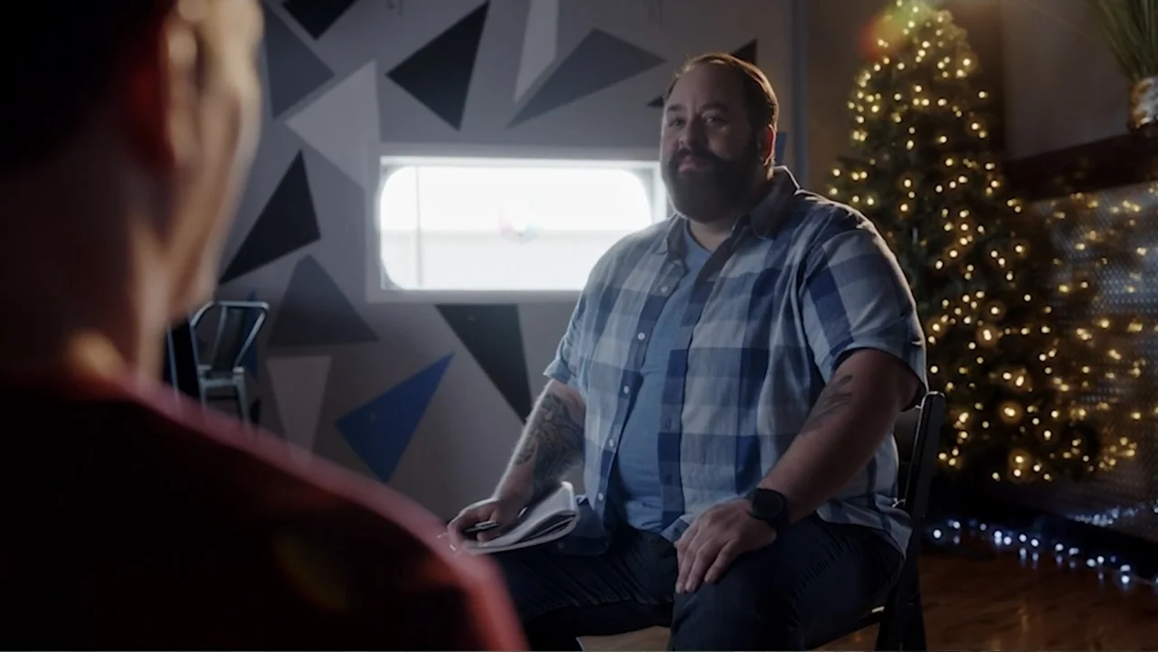 Ask Me to Dance (2022) movie still: medium — A modern office space features a geometric patterned wall and a holiday tree. A bearded man sits in a…