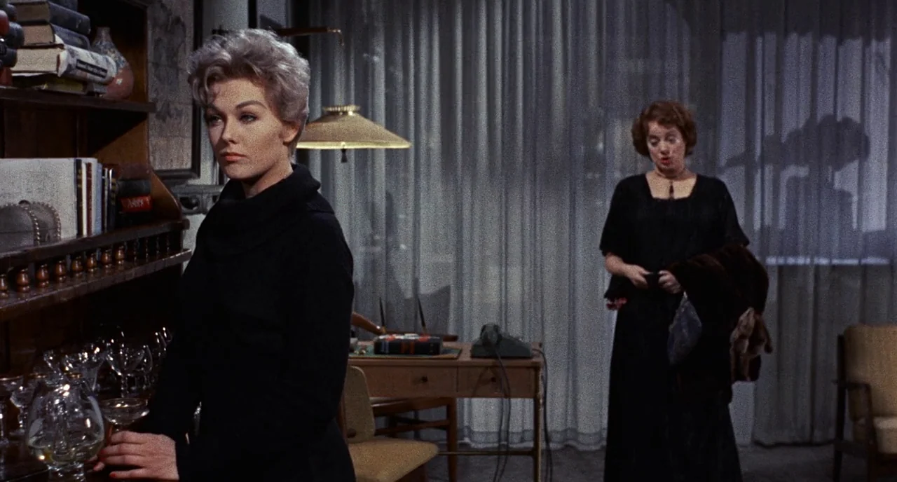 Bell Book and Candle (1958) movie still: medium — A dimly lit room features a bookshelf, a writing desk, and sheer window curtains. One woman stands…