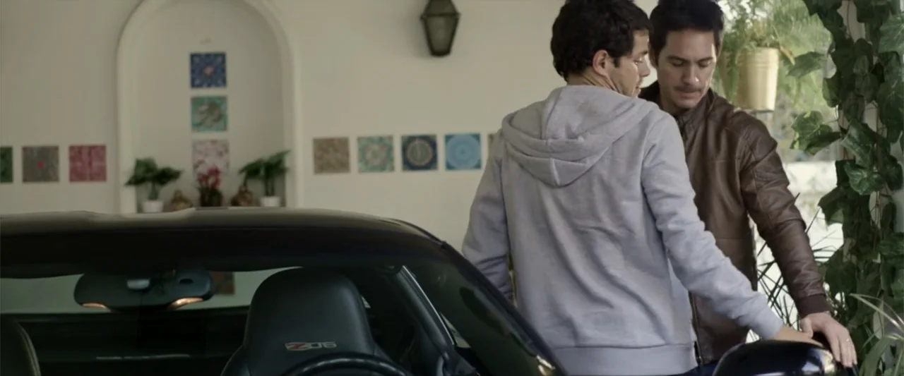 Do It Like An Hombre (2017) movie still: medium — A garage interior features a luxury sports car and wall-mounted decorative tiles. Two men stand…