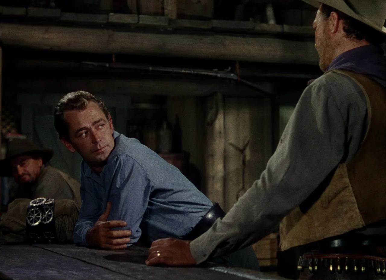 Shane (1953) movie still: medium — A dark wooden interior with rustic timber beams and shelves filled with supplies. Two men stand at a counter in…