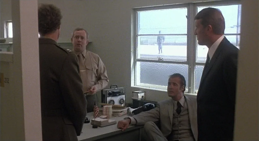Coming Home (1978) movie still: medium — A spartan 1970s room with beige walls, a metal desk, and a large window. Four men in suits and uniforms…