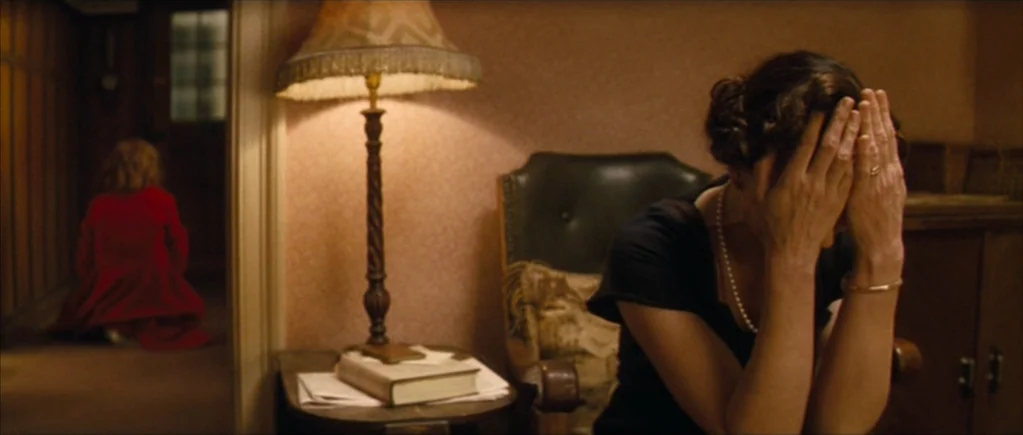 Nowhere Boy movie still: medium — A dimly lit room with wallpapered walls and a wooden table supporting a lamp. A woman covers her face in the…