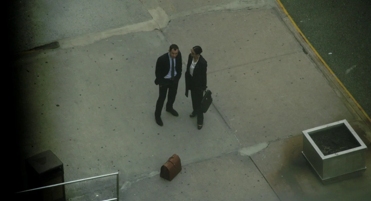 Side Effects (2013) movie still: wide — A wide concrete plaza with a trash bin and a metal planter container nearby. A man and a woman in business…