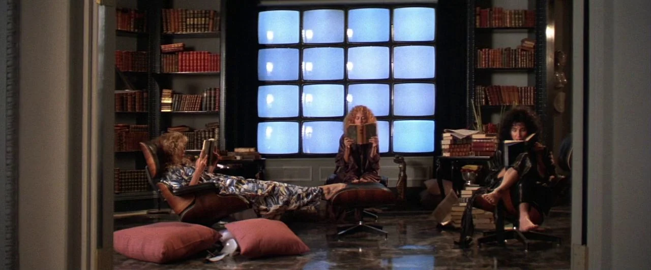 The Witches of Eastwick (1987) movie still: wide — A dark, wood-paneled room features floor-to-ceiling bookshelves and a wall of bright screens.…