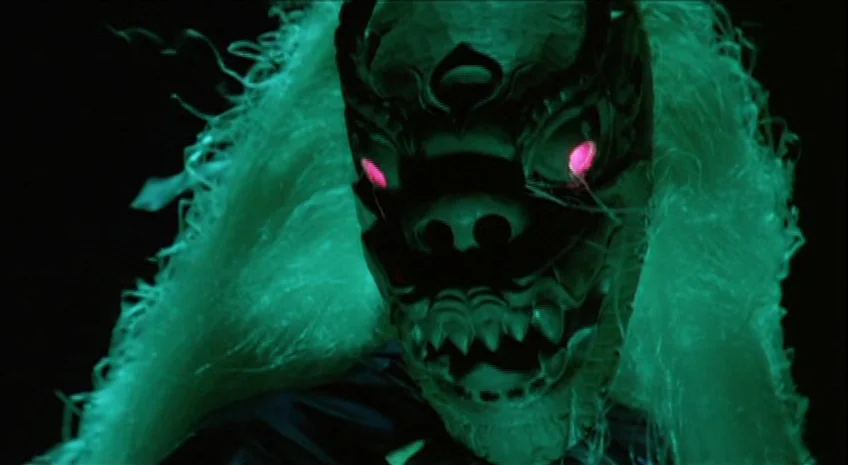 Boxer’s Omen (Mo) (1983) movie still: closeup — A dark environment frames a figure wearing an ornate, creature-like demonic mask. Bright green light…