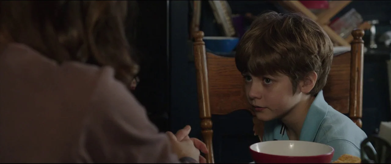 Insidious: Chapter 2 (2013) movie still: medium — A wooden dining table is occupied by a person and a young child. The child watches someone out of…